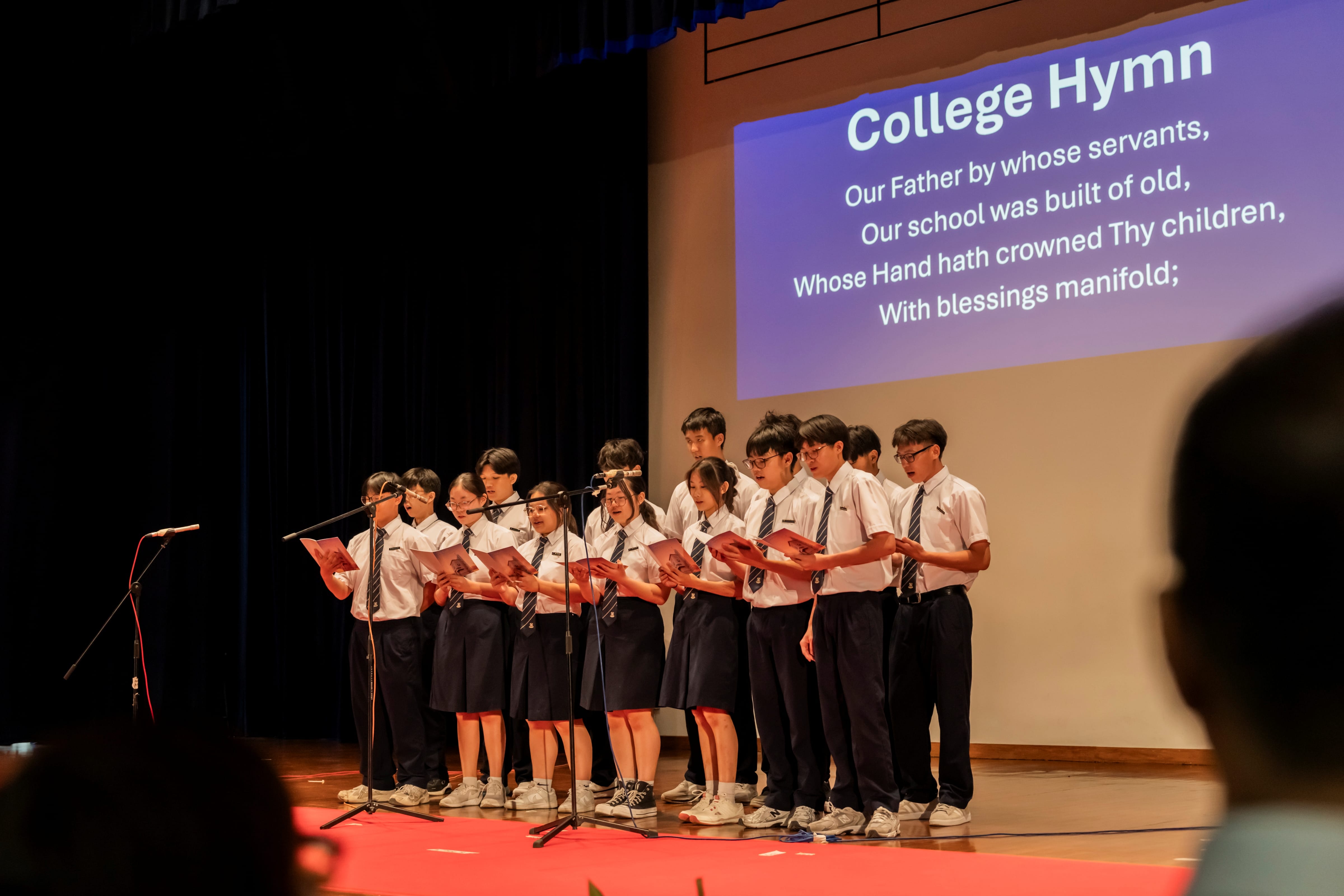 Saints for Christ, a Student Leadership Co-Curricular Activity at SAJC, in school uniforms singing on stage in front of a projected screen displaying the lyrics to the "College Hymn".